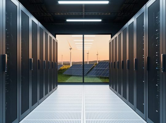 Data center corridor with black server racks under cool blue lighting, and a distant view of wind turbines and solar panels through a glass wall at sunset.